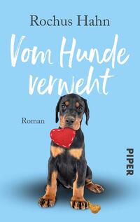 Das Buchcover zeigt einen niedlichen Rottweiler-Welpen, der auf einem hellblauen Hintergrund sitzt. Der Hund hält ein rotes Herzspielzeug im Mund und hat eine verspielte Ausstrahlung. Der Titel „Vom Hunde verweht“ ist in einer freundlichen, handschriftlichen Schriftart in Weiß und gut lesbar platziert. Darunter steht das Wort „Roman“ in einer schlichten Schrift. Unten rechts ist das Logo des Verlags „PIPER“ in Schwarz abgedruckt.