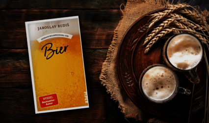 Links liegt das Buch „Gebrauchsanweisung für Bier“ von Jaroslav Rudiš mit gelbem Cover. Rechts stehen zwei Biergläser mit Schaum auf einem braunen Teller, daneben liegen Getreideähren auf einem Jutestoff.