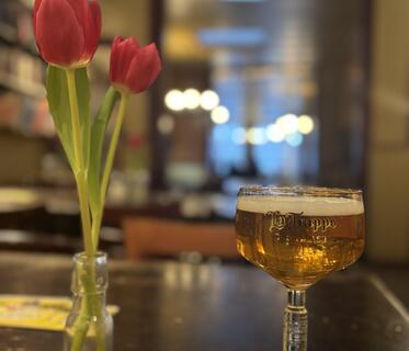 Ein Stielglas mit flachem Kelch, in dem helles Bier eingeschenkt ist und auf dem „La Trappe“ steht, auf einem Holztisch. Daneben stehen zwei rote Tulpen in einer kleinen Vase. Im Hintergrund ist unscharf ein Gastraum mit Lichtern und Regal zu erkennen.