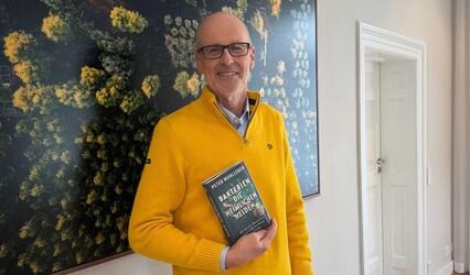 Peter Wohlleben steht mit seinem Buch „Bakterien“ in der Hand vor einer großflächigen Waldfotografie.