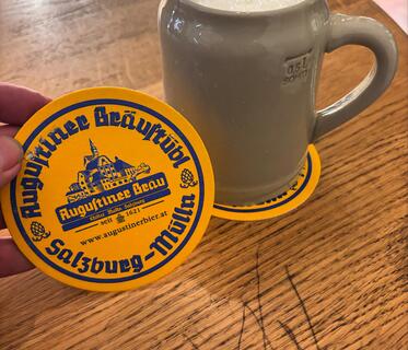 Ein 0,5-Liter-Tonkrug, in dem man weißen Bierschaum sieht, steht auf einem zerkratzten Holztisch. Daneben wird ein gelber Bierdeckel mit der dunkelblauen Aufschrift „Augustiner Bräustübl Salzburg-Mülln“ hochgehalten.