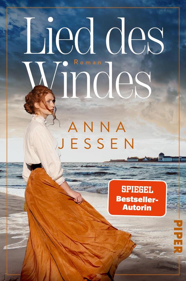 Das Buchcover zeigt eine Frau mit roten Haaren, die in einem weit ausgestellten, orangenen Rock und einer weißen Bluse am Strand steht. Der Himmel ist klar und die Wellen des Meeres sind sichtbar. Über dem Bild prangt der Titel „Lied des Windes“ in eleganten, weißen Buchstaben, während der Autorinnenname „ANNA JESSEN“ darunter in einer goldenen Schriftart erscheint. In der unteren rechten Ecke befindet sich ein rotes Banner mit dem Text „SPIEGEL Bestseller-Autorin“.