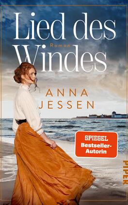 Das Buchcover zeigt eine Frau mit roten Haaren, die in einem weit ausgestellten, orangenen Rock und einer weißen Bluse am Strand steht. Der Himmel ist klar und die Wellen des Meeres sind sichtbar. Über dem Bild prangt der Titel „Lied des Windes“ in eleganten, weißen Buchstaben, während der Autorinnenname „ANNA JESSEN“ darunter in einer goldenen Schriftart erscheint. In der unteren rechten Ecke befindet sich ein rotes Banner mit dem Text „SPIEGEL Bestseller-Autorin“.