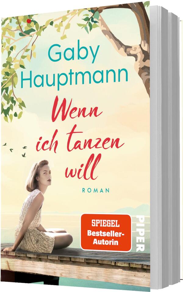 Das Buchcover zeigt eine sommerliche, pastellfarbene Strandlandschaft mit einem Baumast oben rechts und grünen Blättern. Vorne sitzt eine junge Frau in hellem Sommerkleid auf einem Holzsteg, nachdenklich in die Ferne blickend. Oben steht der Autorenname Gaby Hauptmann in türkis, mittig der Titel Wenn ich tanzen will in roter, handschriftlicher Typografie, darunter klein „Roman“. Unten ein orangefarbener Button mit weißer Aufschrift „SPIEGEL Bestseller-Autorin“.
