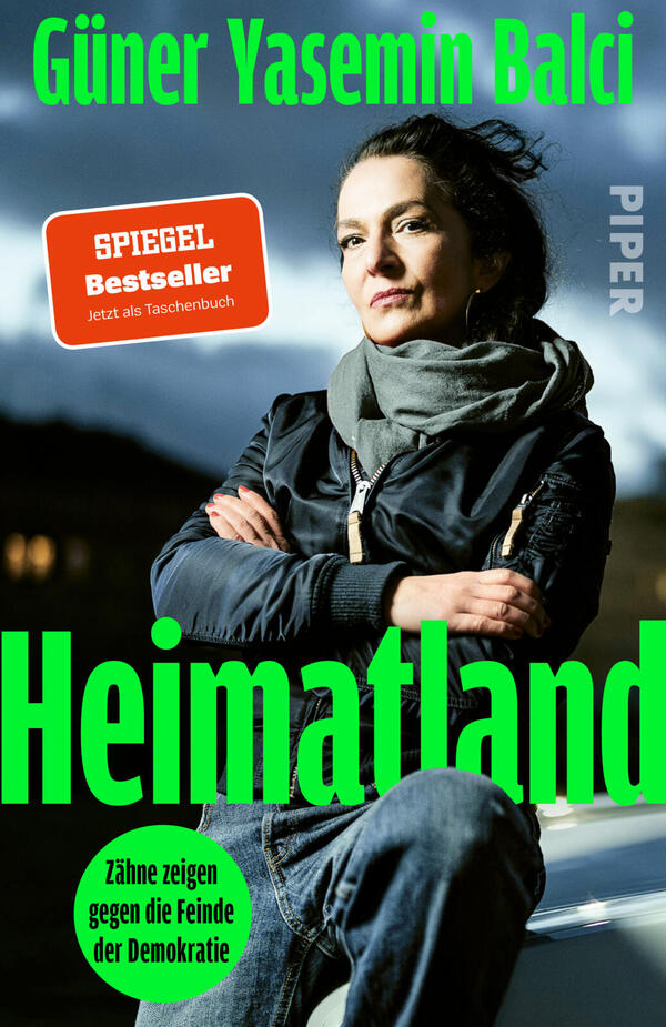 Das Buchcover zeigt eine selbstbewusste Frau mittleren Alters mit dunklen Haaren und Schal vor dramatischem, wolkigem Himmel, frontal stehend mit verschränkten Armen. Dominiert von kräftigem Neongrün für Titel „Heimatland“ und Autorennamen „Güner Yasemin Balcı“, weiße Verlagschrift seitlich. Unten links grüner Kreis mit schwarzem Text „Zähne zeigen gegen die Feinde der Demokratie“. Atmosphäre: ernst, entschlossen.