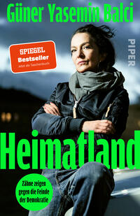 Das Buchcover zeigt eine selbstbewusste Frau mittleren Alters mit dunklen Haaren und Schal vor dramatischem, wolkigem Himmel, frontal stehend mit verschränkten Armen. Dominiert von kräftigem Neongrün für Titel „Heimatland“ und Autorennamen „Güner Yasemin Balcı“, weiße Verlagschrift seitlich. Unten links grüner Kreis mit schwarzem Text „Zähne zeigen gegen die Feinde der Demokratie“. Atmosphäre: ernst, entschlossen.