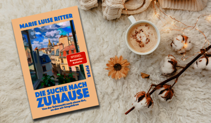 Das Buch „Die Suche nach Zuhause“ liegt neben einer Tasse Kaffee auf einem weißen flauschigenTeppich
