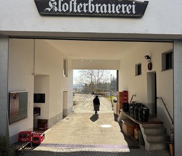Ein gepflasterter Durchgang, über dem der Schriftzug „Klosterbrauerei“ hängt. Im Hintergrund sieht man im Gegenlicht eine Nonne in schwarzem Gewand. Links und rechts an der Wand des Durchgangs stehen Bierkästen, Bierfässer und hängt eine Tafel.