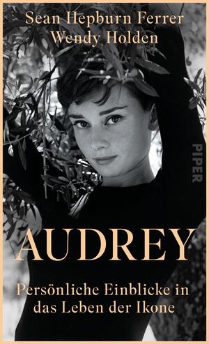 Das Buchcover zeigt eine Schwarz-Weiß-Fotografie von einer Frau mit einem sanften Lächeln, umgeben von grünen Olivenzweigen. Sie trägt einen schwarzen Rollkragenpullover und blickt direkt in die Kamera. Der Titel „AUDREY“ ist in großen, goldenen Buchstaben in der Mitte platziert, darunter steht der Untertitel „Persönliche Einblicke in das Leben der Ikone“ in kleinerer, heller Schrift. Die beiden Autoren, Sean Hepburn Ferrer und Wendy Holden, sind oben in eleganter, weißer Schrift angeführt. Die Atmosphäre ist nostalgisch und stilvoll.
