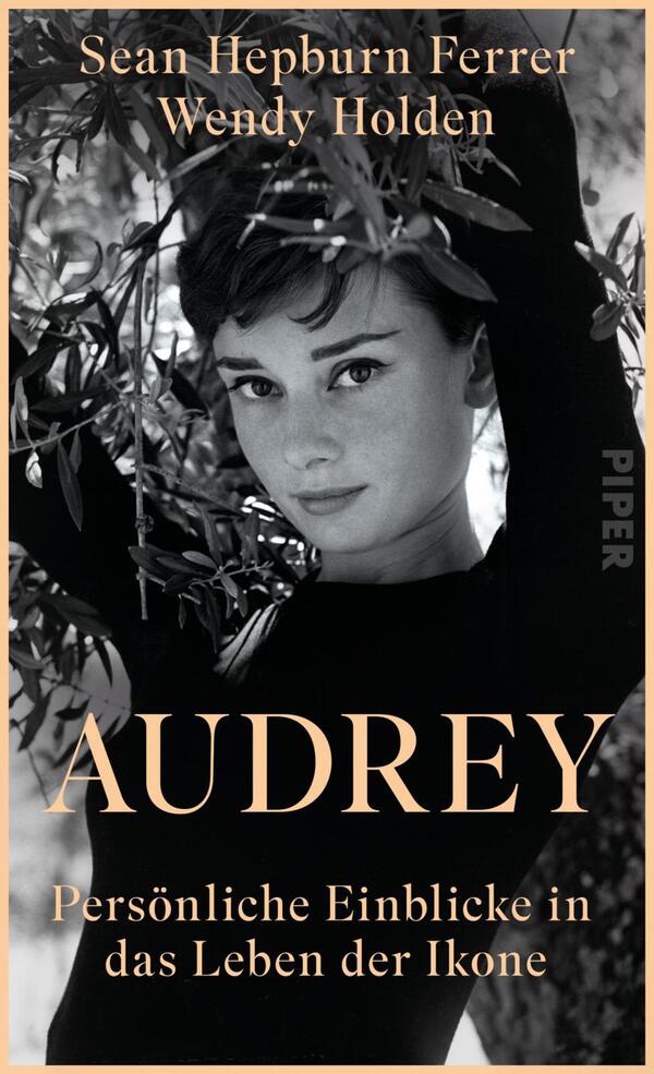 Das Buchcover zeigt eine Schwarz-Weiß-Fotografie von einer Frau mit einem sanften Lächeln, umgeben von grünen Olivenzweigen. Sie trägt einen schwarzen Rollkragenpullover und blickt direkt in die Kamera. Der Titel „AUDREY“ ist in großen, goldenen Buchstaben in der Mitte platziert, darunter steht der Untertitel „Persönliche Einblicke in das Leben der Ikone“ in kleinerer, heller Schrift. Die beiden Autoren, Sean Hepburn Ferrer und Wendy Holden, sind oben in eleganter, weißer Schrift angeführt. Die Atmosphäre ist nostalgisch und stilvoll.