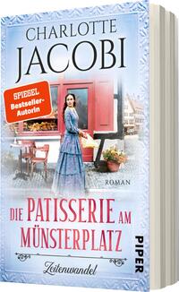 Das Buchcover zeigt eine historische Szenerie mit einer Frau in einem blauen Kleid, die vor einer Patisserie steht. Sie blickt in die Kamera und strahlt Ruhe aus. Im Hintergrund sind eindrucksvolle Hausfassaden und eine einladende Vitrine mit Backwaren zu sehen. Der Titel „Die Patisserie am Münsterplatz“ ist in eleganter Schrift gestaltet und hebt sich deutlich ab. Darüber steht der Name der Autorin „Charlotte Jacobi“ in markanter Schrift. Ein rot umrandetes Siegel trägt die Aufschrift „SPIEGEL-Bestseller-Autorin“.