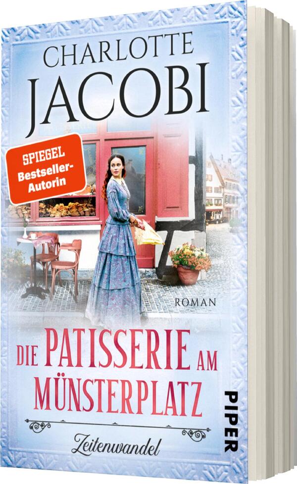 Das Buchcover zeigt eine historische Szenerie mit einer Frau in einem blauen Kleid, die vor einer Patisserie steht. Sie blickt in die Kamera und strahlt Ruhe aus. Im Hintergrund sind eindrucksvolle Hausfassaden und eine einladende Vitrine mit Backwaren zu sehen. Der Titel „Die Patisserie am Münsterplatz“ ist in eleganter Schrift gestaltet und hebt sich deutlich ab. Darüber steht der Name der Autorin „Charlotte Jacobi“ in markanter Schrift. Ein rot umrandetes Siegel trägt die Aufschrift „SPIEGEL-Bestseller-Autorin“.