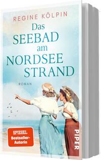 Das Buchcover zeigt ein nostalgisches Strandmotiv mit zwei Frauen in historischer Kleidung, eine mit Sonnenhut und ausgestrecktem Arm, die andere mit Spitzenbluse und Sonnenschirm. Heller Pastellhimmel in Blau und Türkis, sandiger Strand und Möwen. Titel in großen türkisfarbenen Serifbuchstaben „Das SEEBAD am NORDSEE STRAND“, Autorin „Regine Kölpin“, kleiner Zusatz „Roman“. Orangenes Badge unten links mit Text „SPIEGEL Bestseller‑Autorin“.