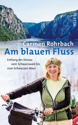 Das Buchcover zeigt eine Frau auf einem Fahrrad, die fröhlich am Ufer eines blauen Flusses posiert. Der Hintergrund besteht aus beeindruckenden, steilen Felsen und einem klaren Himmel. Der Titel „Am blauen Fluss“ steht in großer, blauer Schrift und ist prominent platziert. Darunter ist der Untertitel „Entlang der Donau vom Schwarzwald bis zum Schwarzen Meer“ in kleinerer, schwarzer Schrift. Der Name der Autorin, Carmen Rohrbach, befindet sich ebenfalls oben auf dem Cover.