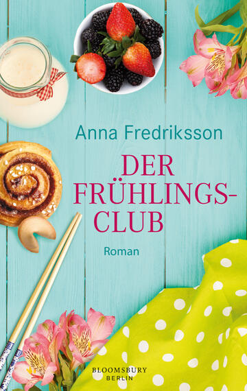 Das Buchcover zeigt einen leicht strukturierten, hellblauen Holzhintergrund. Im oberen Bereich steht in großen, roten Lettern der Titel „DER FRÜHLINGS-CLUB“ und darunter der Schriftzug „Roman“ in kleinerer Schrift. Daneben sind frische Erdbeeren und schwarze Beeren in einer weißen Schüssel abgebildet, während ein Glas Milch, ein Zimtschneckengebäck und ein grüner Stoff mit blühenden, rosa Blumen harmonisch platziert sind. Farbige Essstäbchen ergänzen die Szenerie und verleihen dem Cover eine fröhliche, einladende Atmosphäre.