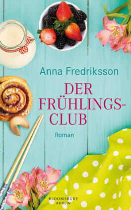 Das Buchcover zeigt einen leicht strukturierten, hellblauen Holzhintergrund. Im oberen Bereich steht in großen, roten Lettern der Titel „DER FRÜHLINGS-CLUB“ und darunter der Schriftzug „Roman“ in kleinerer Schrift. Daneben sind frische Erdbeeren und schwarze Beeren in einer weißen Schüssel abgebildet, während ein Glas Milch, ein Zimtschneckengebäck und ein grüner Stoff mit blühenden, rosa Blumen harmonisch platziert sind. Farbige Essstäbchen ergänzen die Szenerie und verleihen dem Cover eine fröhliche, einladende Atmosphäre.