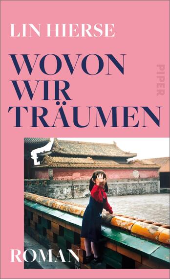 Das Buchcover zeigt einen rosa Hintergrund mit dem Titel „WOVON WIR TRÄUMEN“ in großen, dunkelblauen Buchstaben, platziert auf der rechten Seite. Darunter befindet sich das Wort „Roman“ in kleinerer Schrift. Auf der linken Seite ist ein Bild einer Person zu sehen, die in einem traditionellen Umfeld steht, mit einer roten Jacke und einer nachdenklichen Pose. Die Atmosphäre vermittelt einen Hauch von Nostalgie und träumerischer Reflexion. Oben links steht der Name der Autorin Lin Hierse.