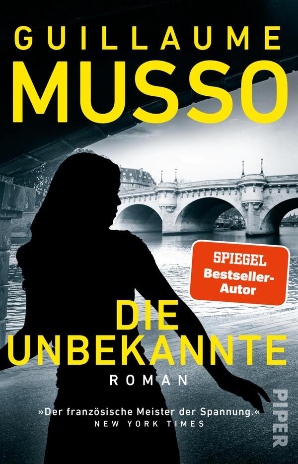 Das Buchcover zeigt eine düstere Silhouette einer Frau vor einer Brücke, die sich über einen Fluss spannt. Der Hintergrund ist in kühlen Grautönen gehalten, während der Titel „DIE UNBEKANNTE“ in leuchtendem Gelb hervorsticht. Darüber steht der Autor „GUILLAUME MUSSO“ ebenfalls in Gelb, und rechts oben befindet sich ein rotes Feld mit dem Text „SPIEGEL Bestseller-Autor“. Die Typografie vermittelt eine spannende und mysteriöse Atmosphäre.