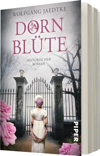 Das Buchcover zeigt eine historische Szene mit einer weiblichen Figur, die von hinten zu sehen ist. Sie trägt ein schlichtes, helles Kleid und einen rosafarbenen Haarreif. Im Hintergrund sind ein großes, offenes Tor und kahle Baumstämme, die eine melancholische Atmosphäre erzeugen. Der Titel „DORNBLÜTE“ ist in satter, pinkfarbener Typografie oben platziert, während darunter in kleinerer Schrift „HISTORISCHER ROMAN“ steht. Rosa Blumen umrahmen das untere Bild.