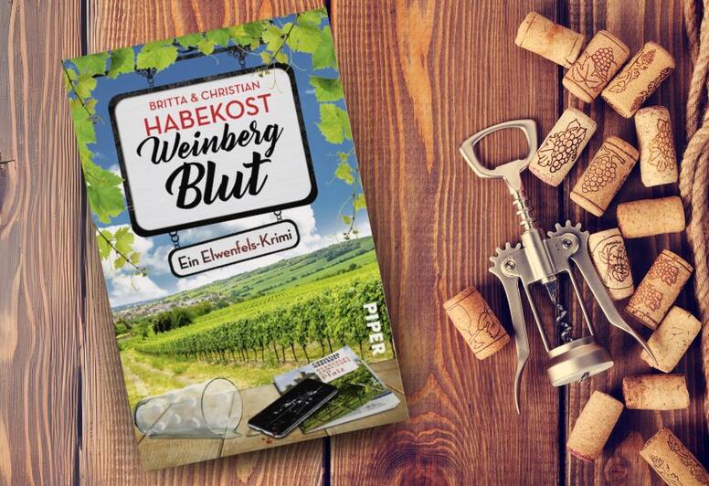 Das Buch 'Weinbergblut' von Britta & Christian Habekost liegt auf einem Holztisch. Neben dem Buch liegen mehrere Weinkorken und ein metallischer Korkenzieher. Das Buchcover zeigt einen Weinberg.