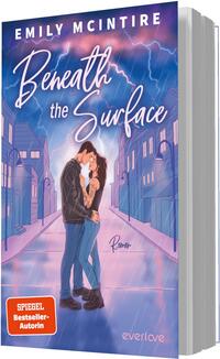 Das Buchcover zeigt ein Paar in inniger Umarmung im Bildmittelpunkt auf einer leeren, perspektivisch geführten Straße zwischen stilisierten Häusern. Der Himmel leuchtet in Violett-, Blau- und Rosa-Tönen mit sichtbaren Blitzen. Titel „Beneath the Surface“ ist groß in weißer, handschriftlicher Schrift gesetzt, der Autorenname EMILY MCINTIRE steht oben in breiter Großschrift. Ein orangener Button trägt den Text „SPIEGEL Bestseller-Autorin“. Atmosphäre: romantisch und elektrisierend.