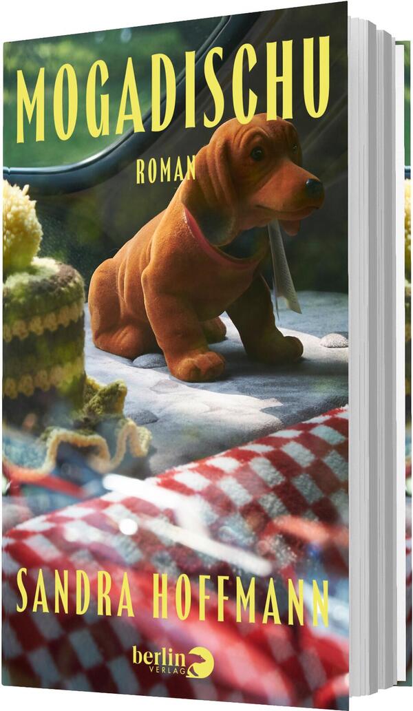 Das Buchcover zeigt ein braunes Plüschtier-Dackel auf einer rot-weiß karierten Picknickdecke, daneben eine gestrickte grün-gelbe Decke, alles in warmem Sonnenlicht vor unscharfem Hintergrund. Der Titel MOGADISCHU steht oben in großen, schmalen gelben Großbuchstaben, unter ihm kleingedruckt „ROMAN“. Unten erscheint der Autorenname Sandra Hoffmann ebenfalls in gelben Großbuchstaben. Stimmung: nostalgisch, heimelig, sonnig.