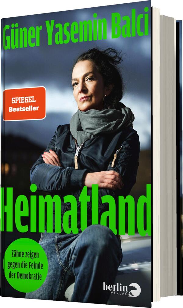 Das Buchcover zeigt eine ernste Frau mit dunklem Haar, die in einer lässigen Jacke und einem Schal gekleidet ist und in die Kamera blickt. Der Hintergrund ist in gedämpften Grautönen gehalten, was die Aufmerksamkeit auf die Figur lenkt. Der Titel „Heimatland“ erscheint in großen, leuchtend grünen Buchstaben im oberen Bereich des Covers. Unterhalb des Titels steht in kleinerer Schrift „Zähne zeigen gegen die Feinde der Demokratie“, ebenfalls in grün. Die Gesamtatmosphäre vermittelt Entschlossenheit und Nachdenklichkeit.