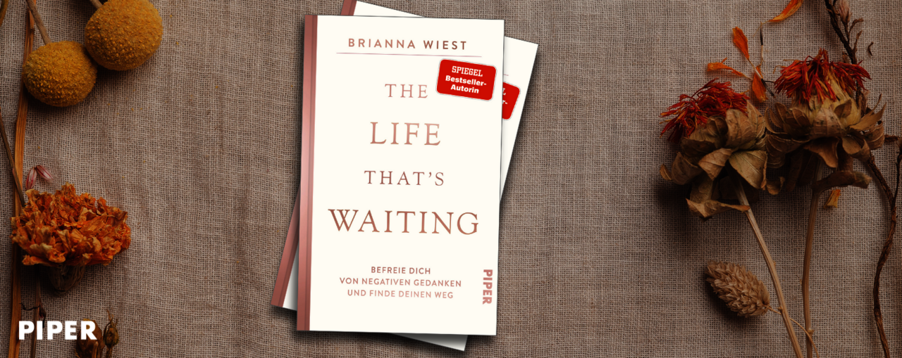 Das Buch "The Life That's Waiting" von Brianna Wiest liegt auf einem beigen Stoff. Daneben sind getrocknete Blumen und gelbe Kugelblüten arrangiert.
