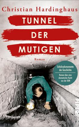 Das Buchcover zeigt einen dunklen Tunnel, in dem ein Mann mit einer Taschenlampe kriechend abgebildet ist. Die Farben sind überwiegend grau und schwarz, mit auffälligen roten Akzenten. Der Titel „TUNNEL DER MUTIGEN“ ist in großen, weißen Buchstaben auf rotem Hintergrund platziert. Darunter steht „Roman“ in kleinerer Schrift. Ein Rundlogo oben rechts trägt die Aufschrift „Schicksalsmomente der Geschichte“ und beschreibt den Inhalt des Buches.