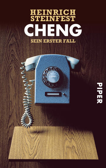 Das Buchcover zeigt ein blaues Wählscheibentelefon auf einem hölzernen Untergrund. Im Hintergrund ist eine dunkelbraune Holzstruktur erkennbar, die eine warme Atmosphäre vermittelt. Der Titel „CHENG“ steht in großen, weißen Buchstaben hervor und ergänzt den Untertitel „sein erster Fall“ in kleinerer Schrift. Der Autorenname „HEINRICH STEINFEST“ befindet sich am oberen Rand in hellen Buchstaben. Das Verlagslogo „PIPER“ ist in der unteren rechten Ecke platziert.