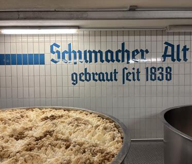 Ein großer Behälter mit schäumendem Bier in offener Gärung. Im Hintergrund ist eine weiß geflieste Wand mit der dunkelblauen Aufschrift „Schumacher Alt – gebraut seit 1838“ zu sehen.