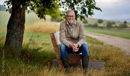 Peter Wohlleben sitzt auf einer Bank vor landschaftlichem Hintergrund.