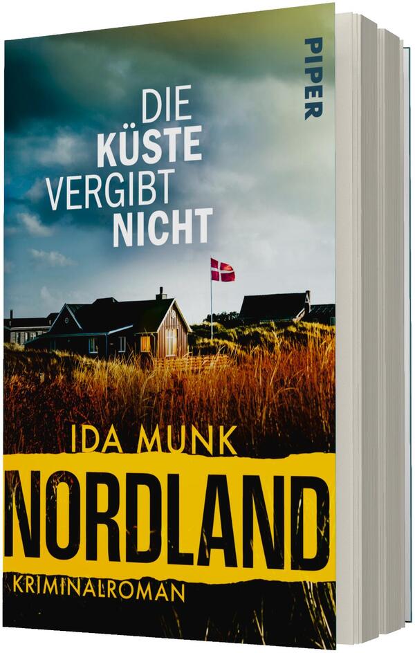 Das Buchcover zeigt eine dänische Küstenlandschaft mit niedrigen Häusern hinter hohen, goldbraunen Dünenhalmen und einer wehenden dänischen Flagge vor dramatisch wolkigem, blau-grünem Himmel. Oben steht der Titel „Die Küste vergibt nicht“ in großen weißen Versalien; darunter der Autorenname Ida Munk in gelber Schrift. Unten ein breiter gelber Streifen trägt den großen schwarzen Schriftzug „NORDLAND“ und klein „Kriminalroman“. Atmosphäre: düster und bedrohlich.