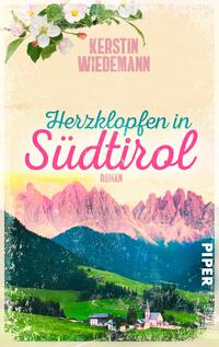Das Buchcover zeigt einen pastellfarbenen Himmel auf beigem Hintergrund, oben Blütenzweige, darunter in rosafarbenen Großbuchstaben der Autorenname „Kerstin Wiedemann“. Titel in schwungvoller Typografie: „Herzklopfen in“ in türkisem Kursiv, „Südtirol“ groß und pink, darunter kleiner Schriftzug „Roman“. Im unteren Bereich eine grüne Almlandschaft mit Dorfkirche und rosa beleuchteten Dolomiten im Abendlicht. Stimmung: romantisch, warm, nostalgisch.