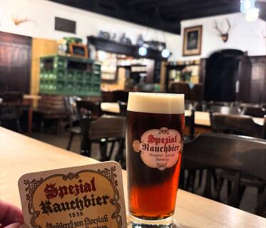 Ein Glas mit der Aufschrift „Spezial Rauchbier“ steht auf einem Holztisch. Neben dem Glas wird der passende Bierdeckel hochgehalten. Im Hintergrund ist eine rustikale Gaststätte mit Holzmöbeln und einem grün gekachelten Ofen sichtbar. 