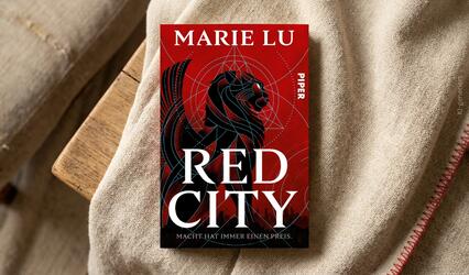 „Red City“ von Marie Lu liegt auf hellem Stoff auf einem Holztisch.