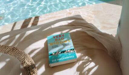Am Rand eines blauen Pools liegt das Buch „Very Bad Widow“. Schatten von Palmenblättern fallen auf die Szene.
