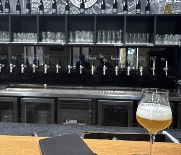 Ein Stielglas mit hellem Bier und ein schwarzes Notizbuch liegen auf einer Holztheke. Im Hintergrund sind eine Reihe silberner Zapfhähne zu sehen, darüber Gläser und Flaschen in einem schwarzen Regal sowie an der mit Kreide bemalten schwarzen Wand darüber das Logo der Freien Brau Union Bremen.