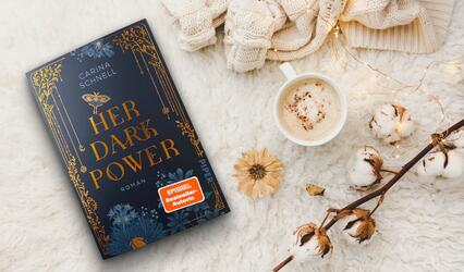 „Her Dark Power“ von Carina Schnell liegt auf einem flauschigen Untergrund. Daneben eine Tasse mit Cappuccino, eine Baumwollast sowie ein gestrickter Pulli.