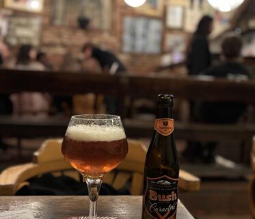 Ein hohes Glas mit dunkelbraunem Bier steht neben einer dunklen „Bush Caractère“-Flasche auf einem Holztisch. Im Hintergrund ist verschwommen eine belgische Bar mit Backsteinwänden, Bildern und runden Hängelampen zu sehen.