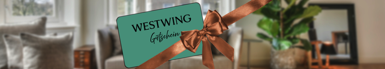 Eine Westwing-Gutscheinkarte steckt unter einer goldenen Schleife, im Hintergrund ist unscharf eine helle, modern eingerichtete Wohnung zu sehen