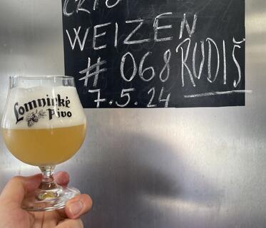 Ein bauchiges Glas mit hellem Bier und einer Schaumkrone wird vor eine schwarze Tafel gehalten. Auf der Tafel steht mit weißer Kreide unter anderem geschrieben: „Weizen #068 Rudiš“ und das Datum „5.4.24“.
