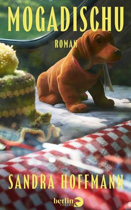 Das Buchcover zeigt einen braunen Plüschdackel, der auf einer gepolsterten Decke sitzt; im Vordergrund ein rot‑weiß kariertes Picknicktuch und links ein gehäkeltes, gelb‑grünes Stoffstück. Der Hintergrund ist unscharf-grün, warm beleuchtet. Der Titel „MOGADISCHU“ und das Wort „ROMAN“ erscheinen oben in großen gelben Großbuchstaben, der Autorenname „SANDRA HOFFMANN“ steht unten in gleicher Typografie. Atmosphäre nüchtern, leicht nostalgisch.