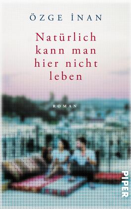 Das Buchcover zeigt eine unscharfe, verträumte Szenerie mit zwei Figuren, die auf einem Teppich sitzen und den Blick auf eine Stadtlandschaft richten. Der Hintergrund ist in sanften Rosa- und Blautönen gehalten, was eine melancholische Atmosphäre vermittelt. Der Titel „Natürlich kann man hier nicht leben“ ist zentral in kräftigem Rot platziert, während der Autorennamen „Özge İnan“ in einem eleganten dunklen Blau oben steht. Der Untertitel „Roman“ ist dezent in Kleinbuchstaben abgedruckt.