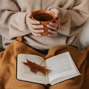 Eine Frau hält eine Teetasse mit beiden Händen vor ihrem Körper. Auf ihrem mit einer Decke bedecktem Schoß liegt ein aufgeschlagenes Buch.
