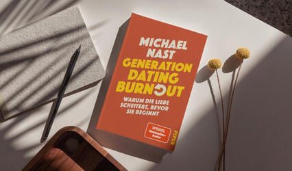 „Generation Dating Burnout“ liegt auf einem hellen Tisch. Links liegt ein graues Notizbuch mit Stift. Rechts liegen drei gelbe Blüten mit langen Stielen. Sonnenlicht wirft Schatten auf die Fläche.