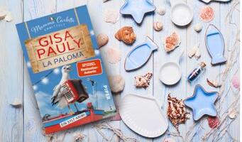 Das Buch 'La Paloma' von Gisa Pauly liegt auf einem hellen Holztisch. Es ist von Muscheln, kleinen Dekorationsformen in Form von Fischen und Sternen umgeben.