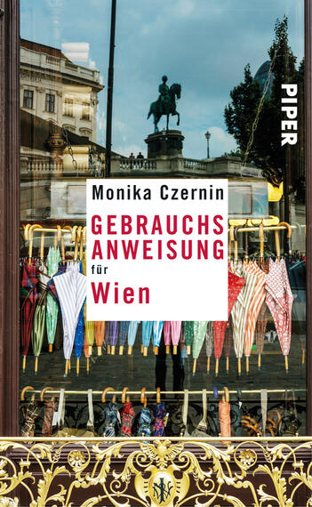 Das Buchcover zeigt eine Schaufensteransicht mit einem bunten Sortiment an Regenschirmen in verschiedenen Farben, darunter gestreifte und einfarbige Modelle. Über den Schirmen steht in großen roten Buchstaben der Titel „GEBRAUCHSANWEISUNG für Wien“. Darunter ist der Name der Autorin Monika Czernin in kleinerer Schrift platziert. Im Hintergrund ist ein neoklassizistisches Gebäude und eine Statue zu erkennen, die eine städtische Atmosphäre schaffen. Der Verlagsname „PIPER“ erscheint oben rechts in weißer Schrift.