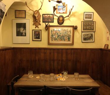 Kleine Nische in einer Kneipe, die mit Holz vertäfelt ist und in der ein rustikaler Tisch mit fünf leeren Bierkrügen steht. Darüber wölbt sich eine gelbe Decke. An den Wänden hängen Fotos und Geweihe.