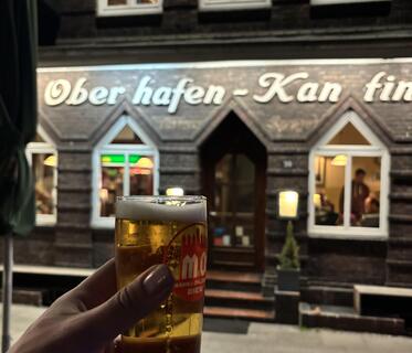 Ein schmales Glas mit hellem Bier wird vor der beleuchteten Oberhafen-Kantine in Hamburg gehalten. Die Fassade des Gebäudes ist dunkel verkleidet und zeigt in Weiß den Schriftzug der Kantine. Tür und Fenster verlaufen nach oben zu spitzen Dreiecken.